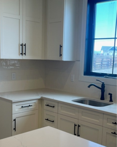 Bright corner kitchen with white cabinets, a marble countertop, built-in sink and faucet, and a window with blue frame showing an outdoor view.